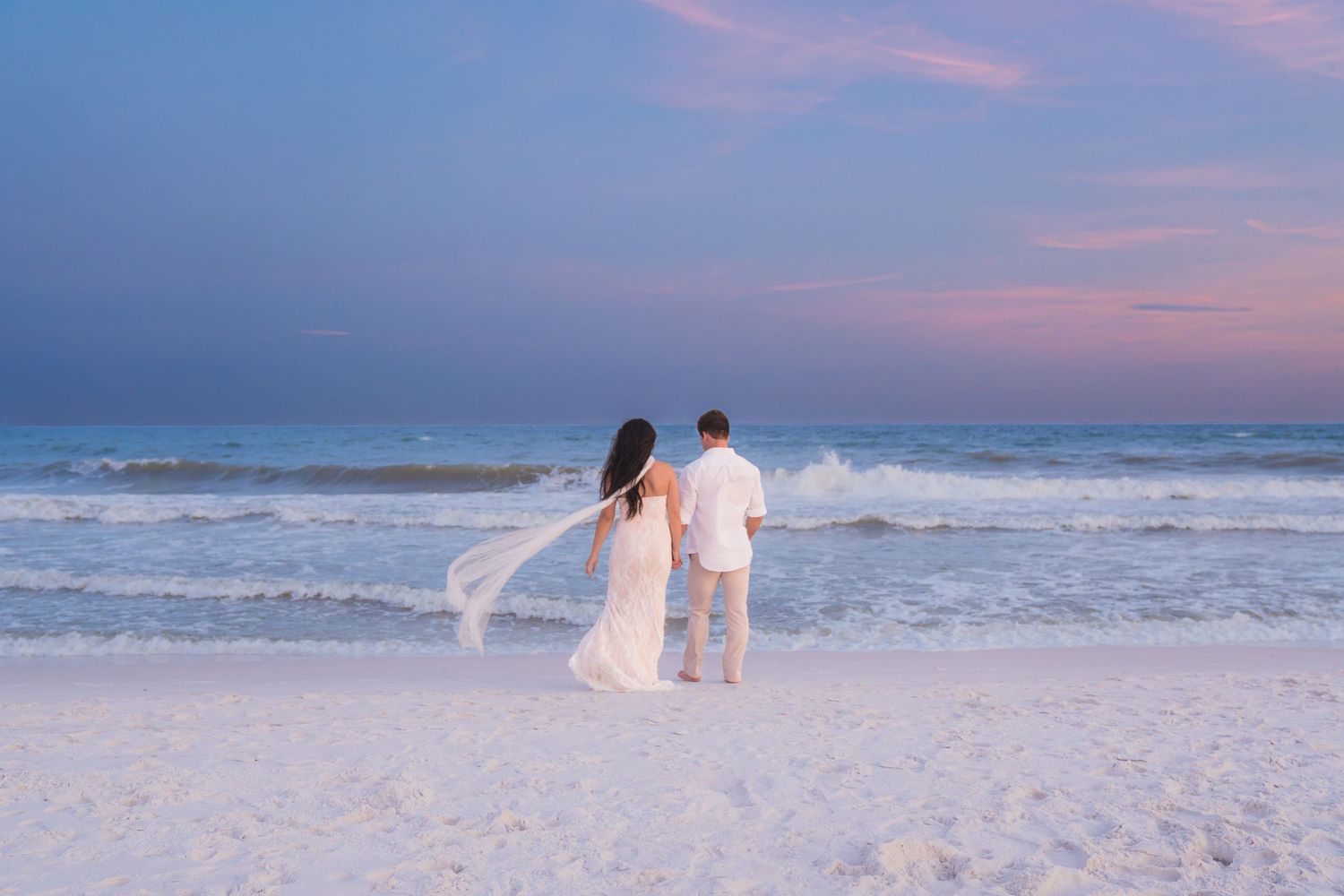 Couple in white attire standing on a sandy beach at sunset with pink and purple sky and ocean waves.