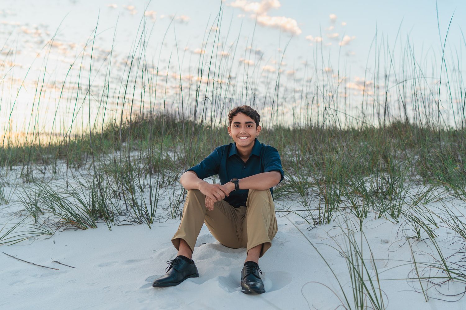 Young man smiling on white sand dunes with sea oats at sunset beach.
