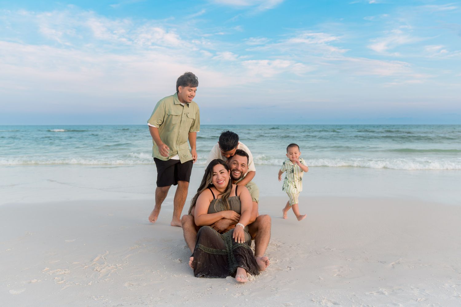 Happy family posing together on a white sandy beach with two children and calm turquoise ocean in background.