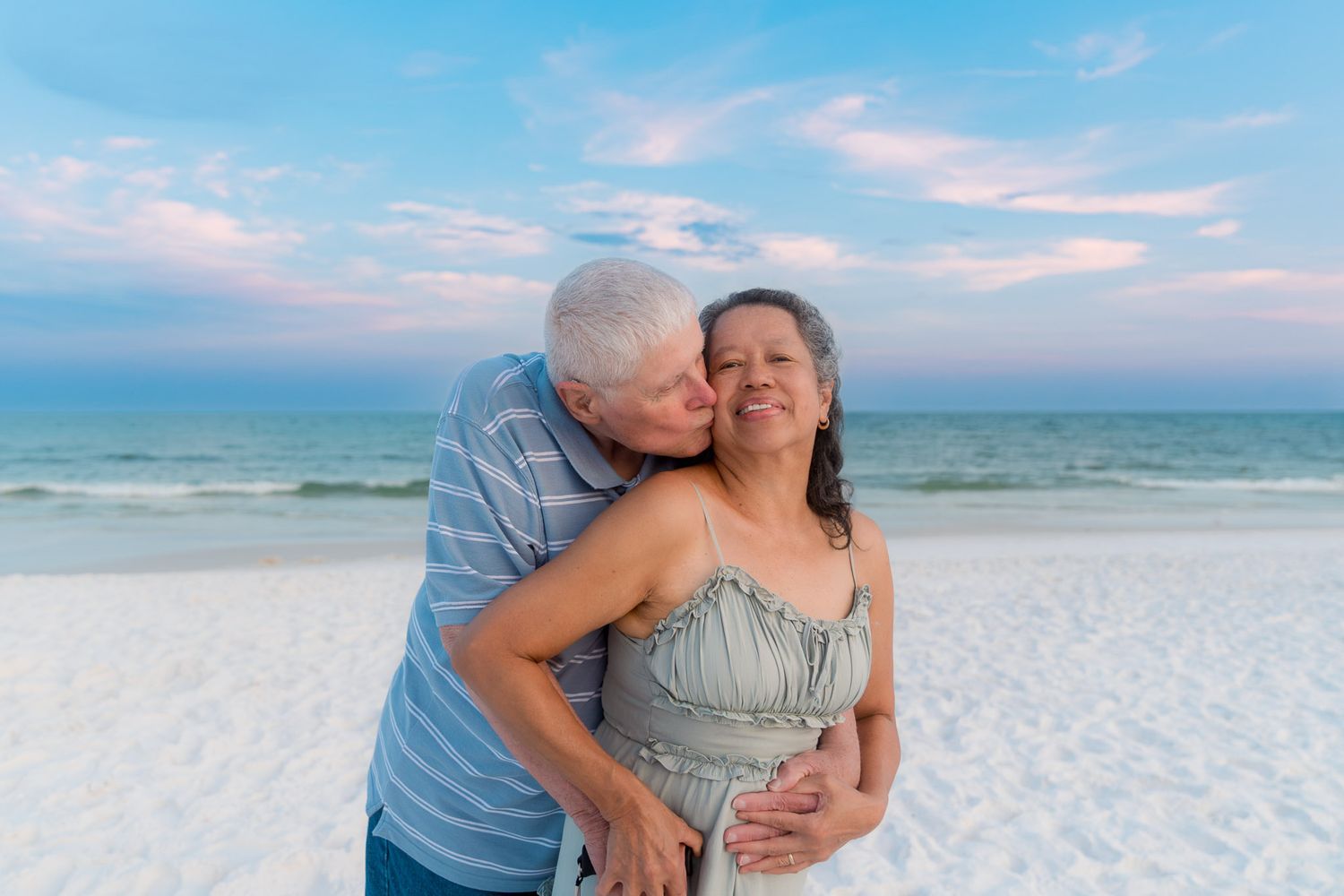 Elderly couple sharing a romantic kiss on a white sand beach at sunset with pastel skies and ocean waves.