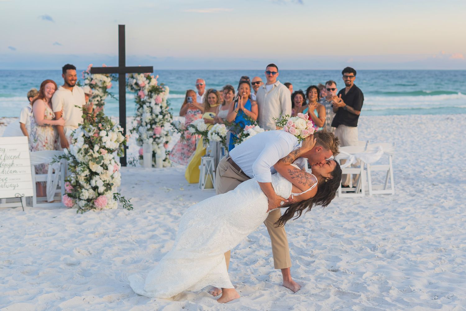 Groom dips bride in romantic beach wedding kiss with guests cheering near floral cross altar at sunset.