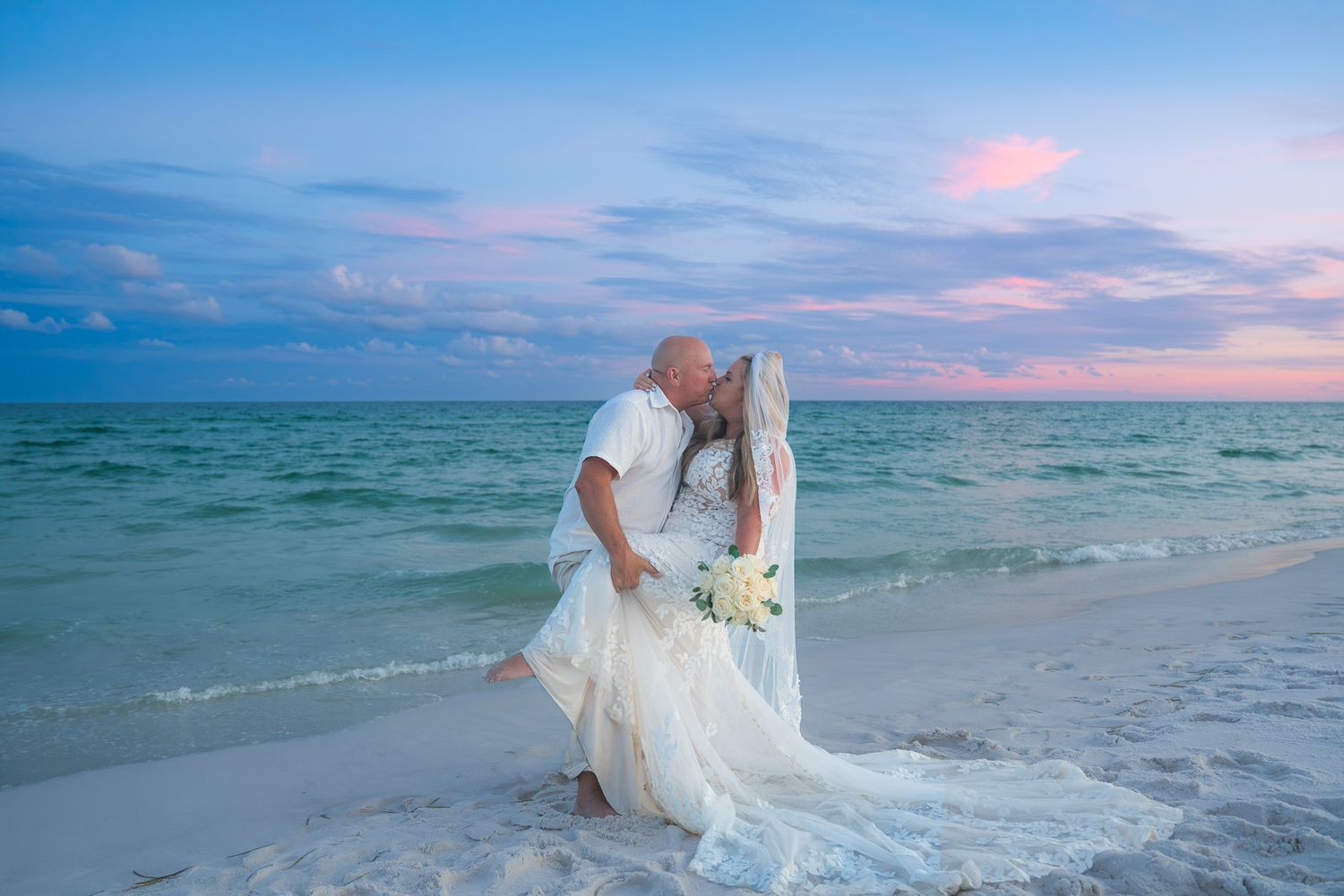 Bride and groom share a romantic kiss on a sandy beach at sunset, with turquoise ocean waves behind them.