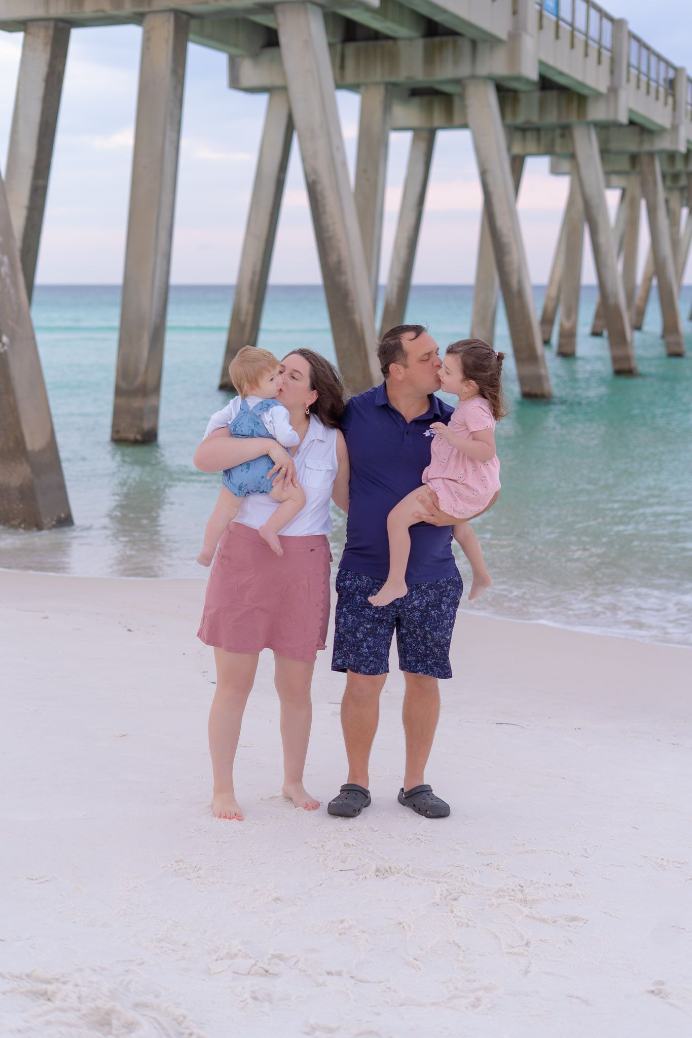 A happy family of four shares a kiss on a white sand beach near a wooden pier at sunset.
