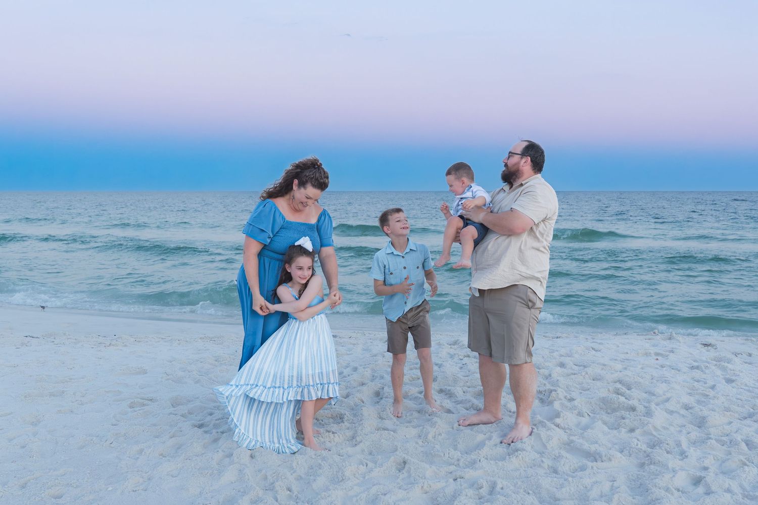 Family of five posing on sandy beach at sunset, parents with three young children, calm ocean waves in background.