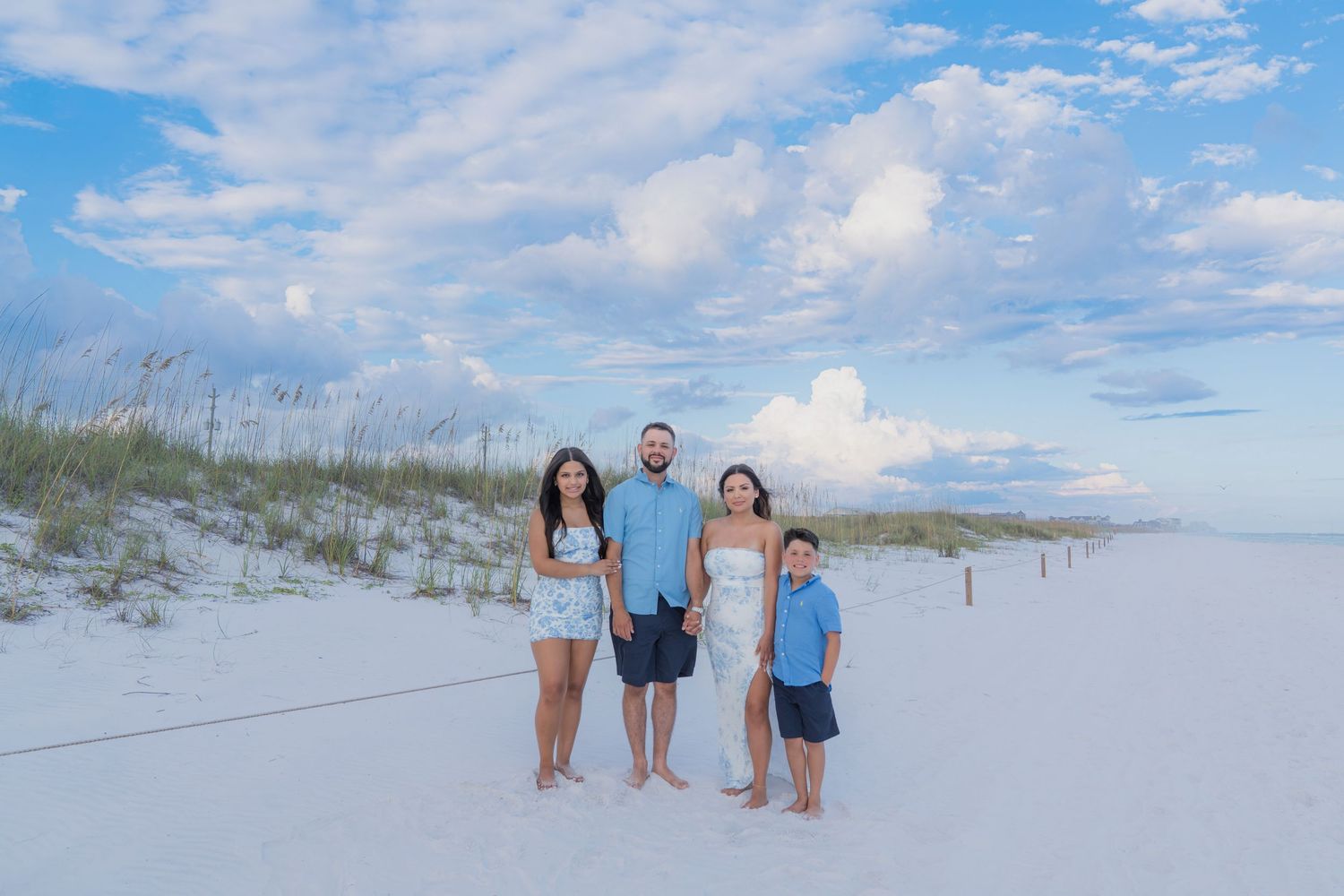 Family of four posing on white sand beach with blue sky and clouds behind them.