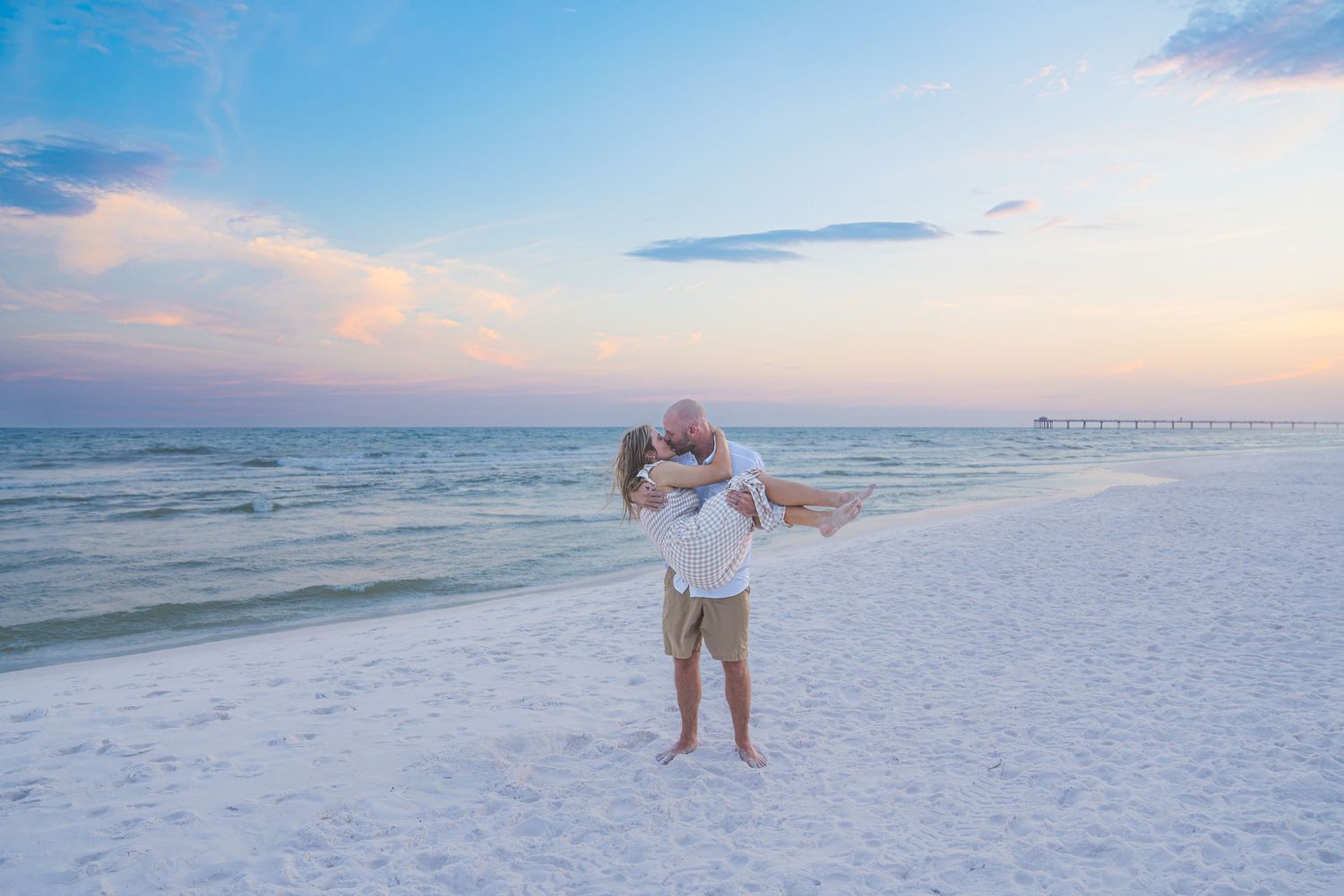 Couple embracing on white sand beach at sunset, man lifting woman in romantic pose with ocean and colorful sky behind them.