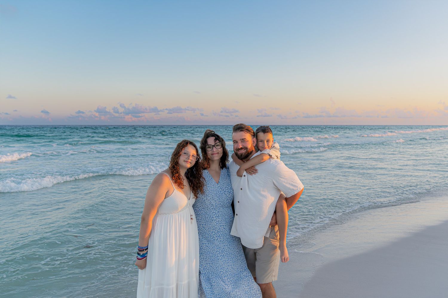 Family of four smiling together on a white sand beach at sunset with turquoise ocean waves behind them.