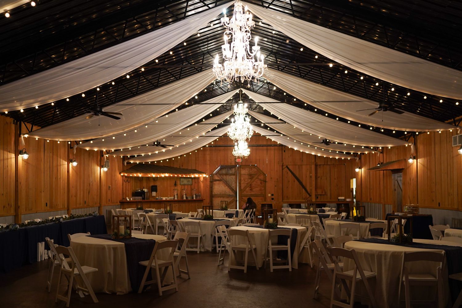 Elegant barn wedding reception venue with string lights, white draping, chandelier, and round tables with white chairs.