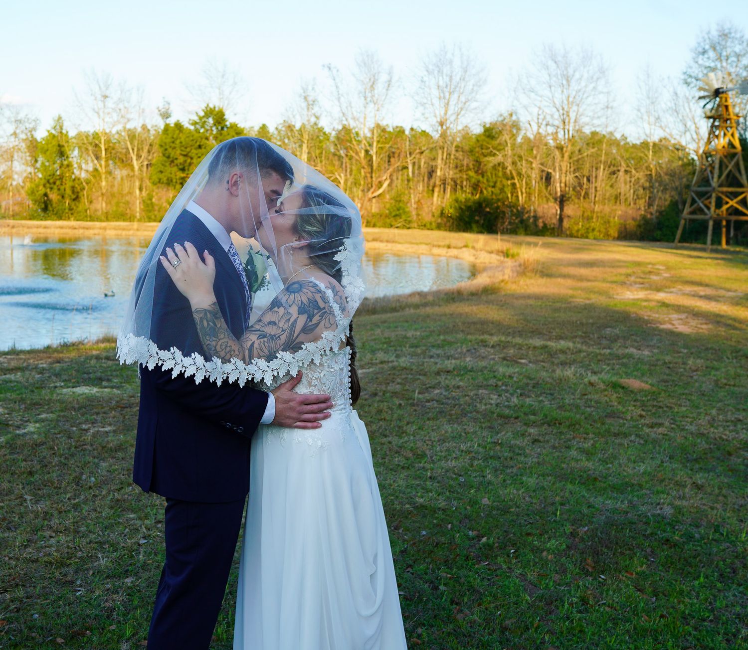 Bride and groom sharing a romantic kiss under a lace veil by a serene pond at their outdoor wedding.