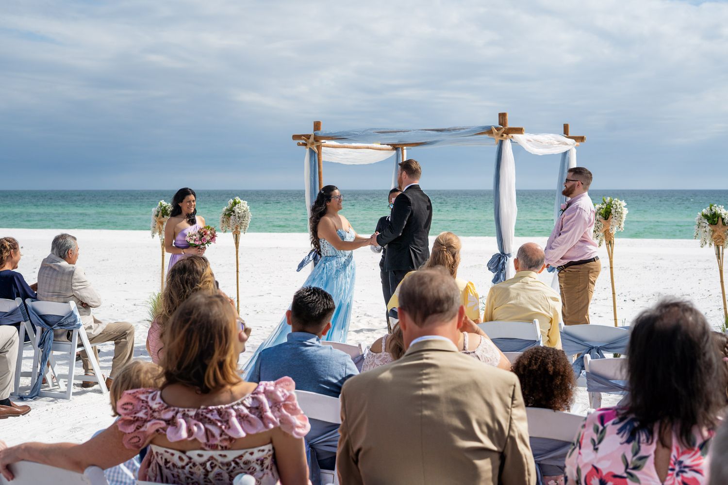Beach wedding ceremony with bride in blue gown and guests seated on white sand.