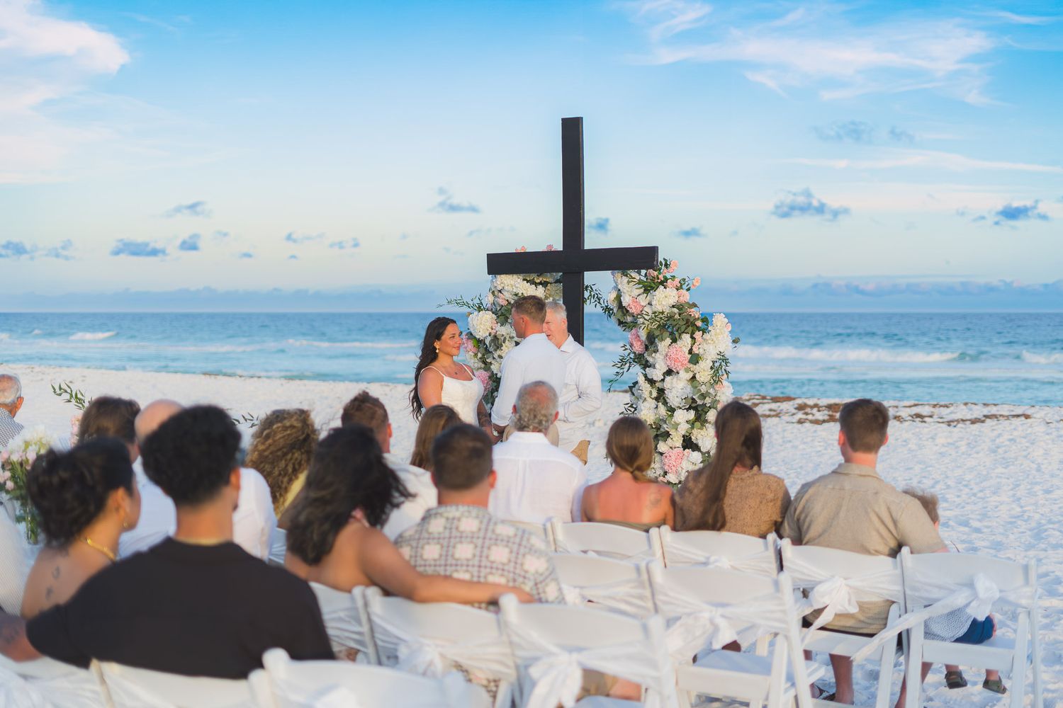 Guests seated at an outdoor beach wedding ceremony with a floral cross altar and ocean backdrop.