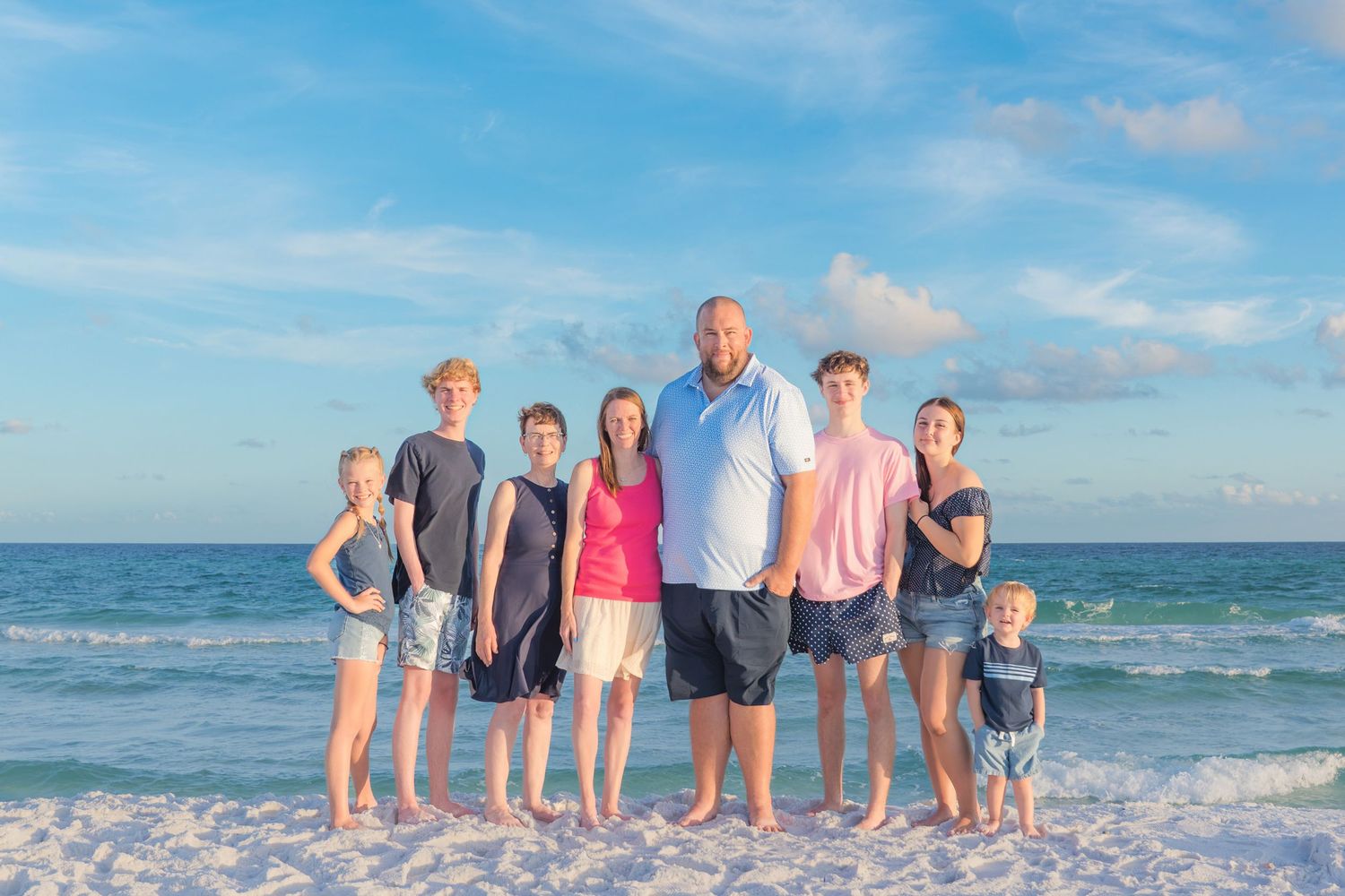 Family of seven posing together on a white sand beach with turquoise ocean waves and blue sky behind them.