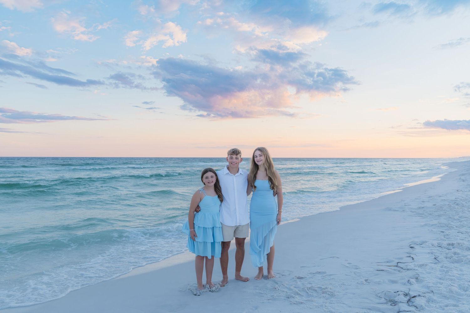 Three people in blue and white outfits pose on a white sand beach at sunset with turquoise waves behind them.