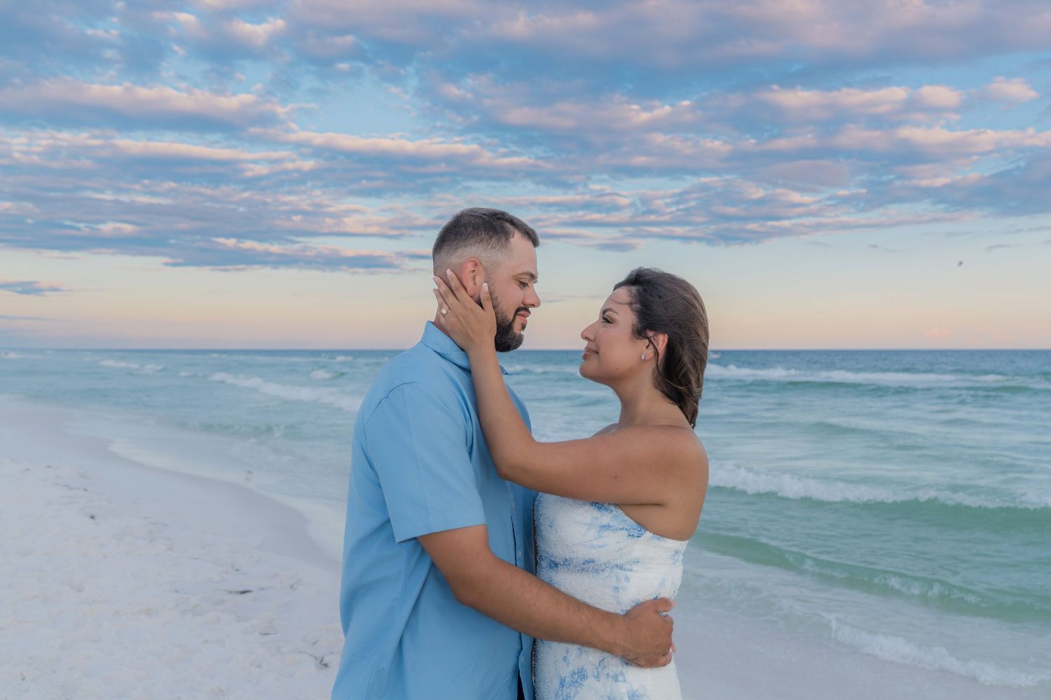 Couple embracing on a white sand beach at sunset with turquoise water and pastel sky behind them.