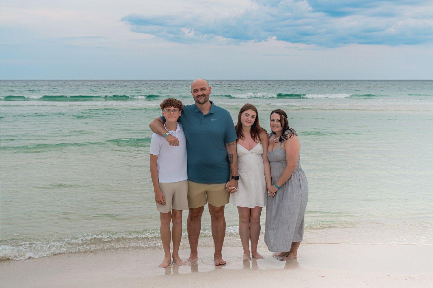 Family of four smiling together on a white sandy beach with turquoise ocean waves in the background.