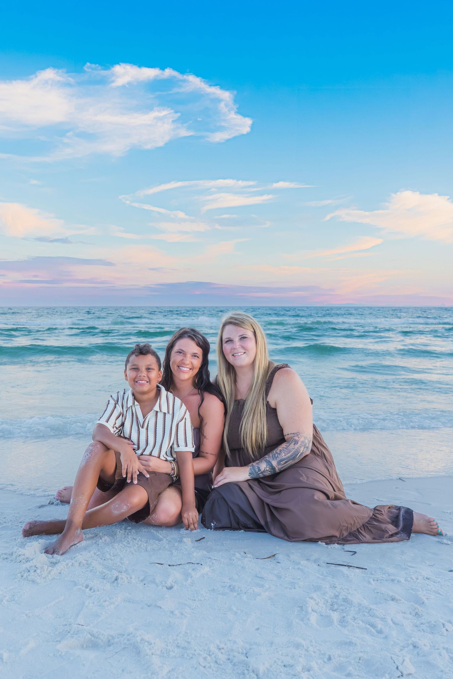 Three people sitting together on a white sand beach at sunset with turquoise ocean waves in the background.