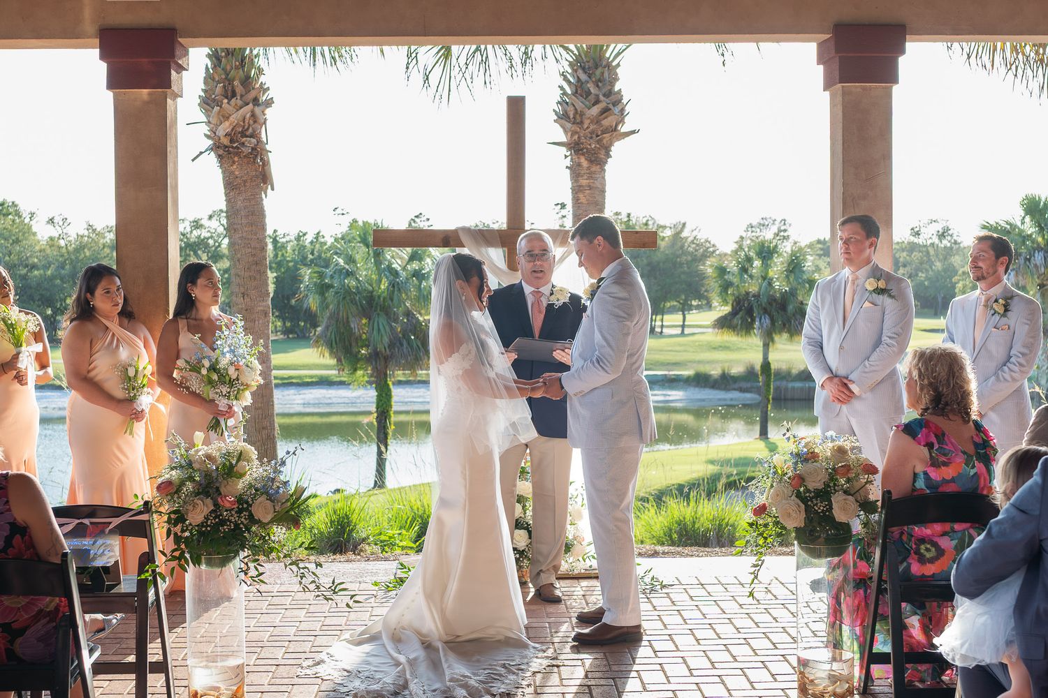 Bride and groom exchange vows under pavilion with wedding party and guests at outdoor waterfront ceremony.