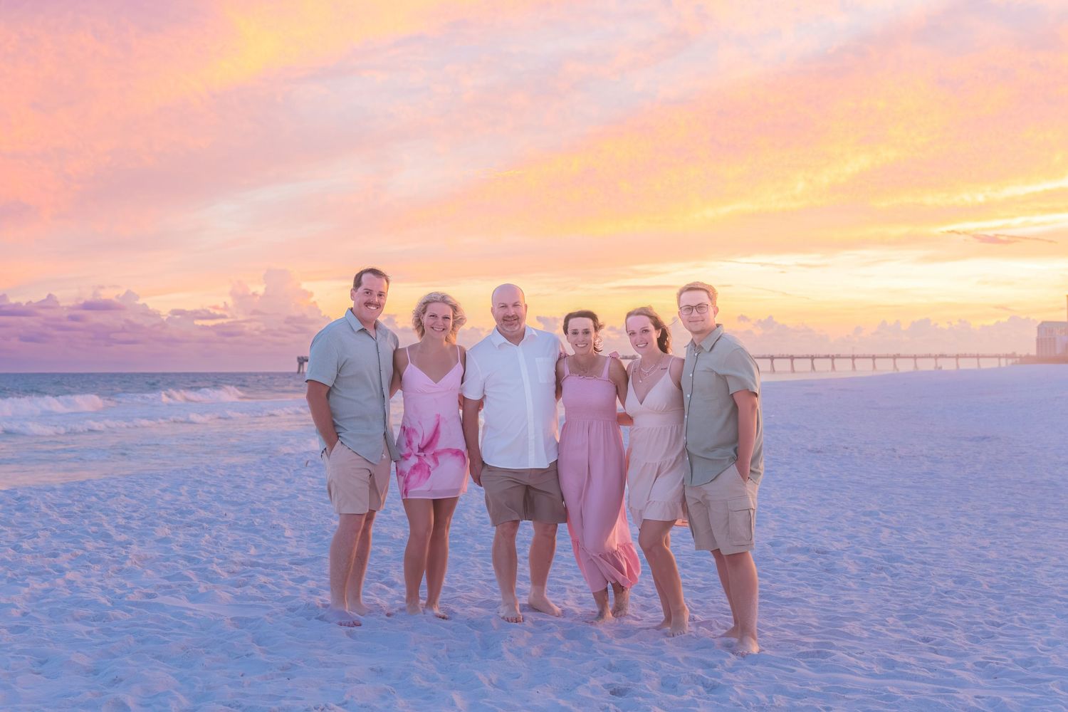 Family of six smiling together on beach during vibrant pink and orange sunset.