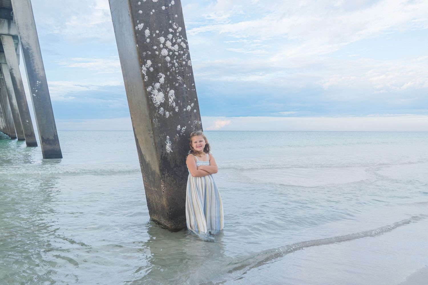 Young girl in blue dress standing between pier pillars in ocean water.
