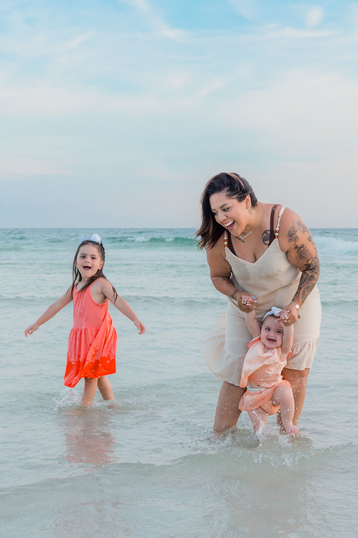 A tattooed mom helps her baby walk in ocean waves while her young daughter in a coral dress plays nearby at the beach.