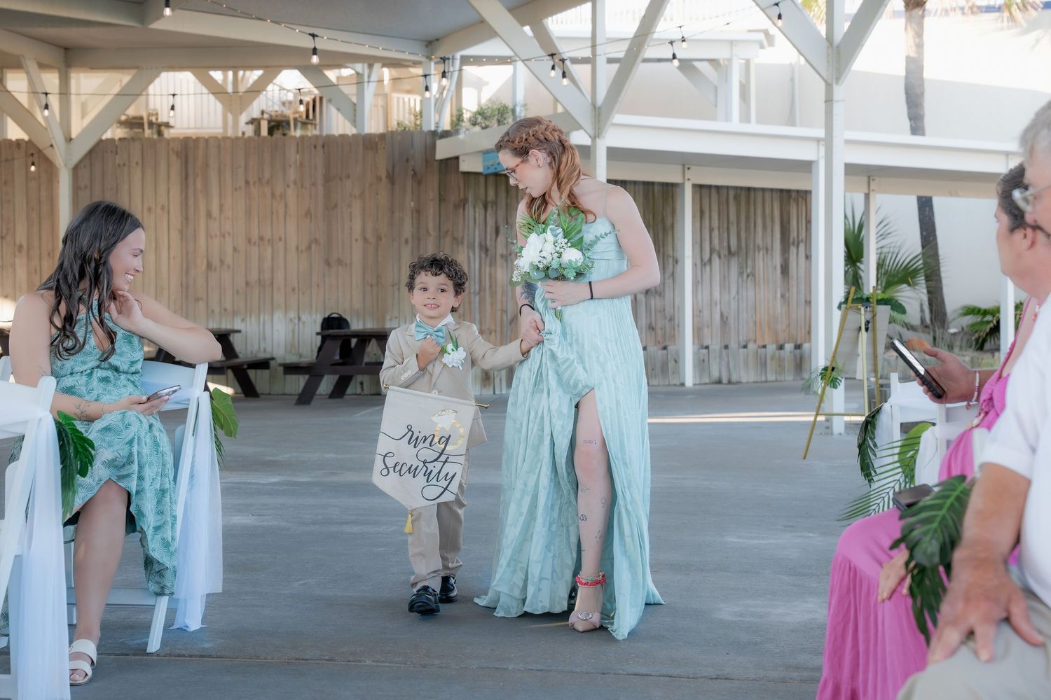 Bridesmaid in mint green dress walking with young ring bearer carrying burlap sign down aisle.