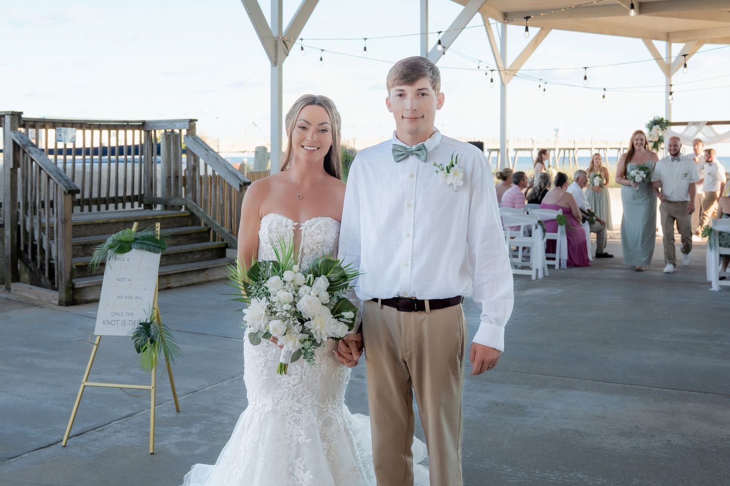 Bride in mermaid gown holding white bouquet stands with groom in khakis and bow tie at outdoor waterfront wedding venue.