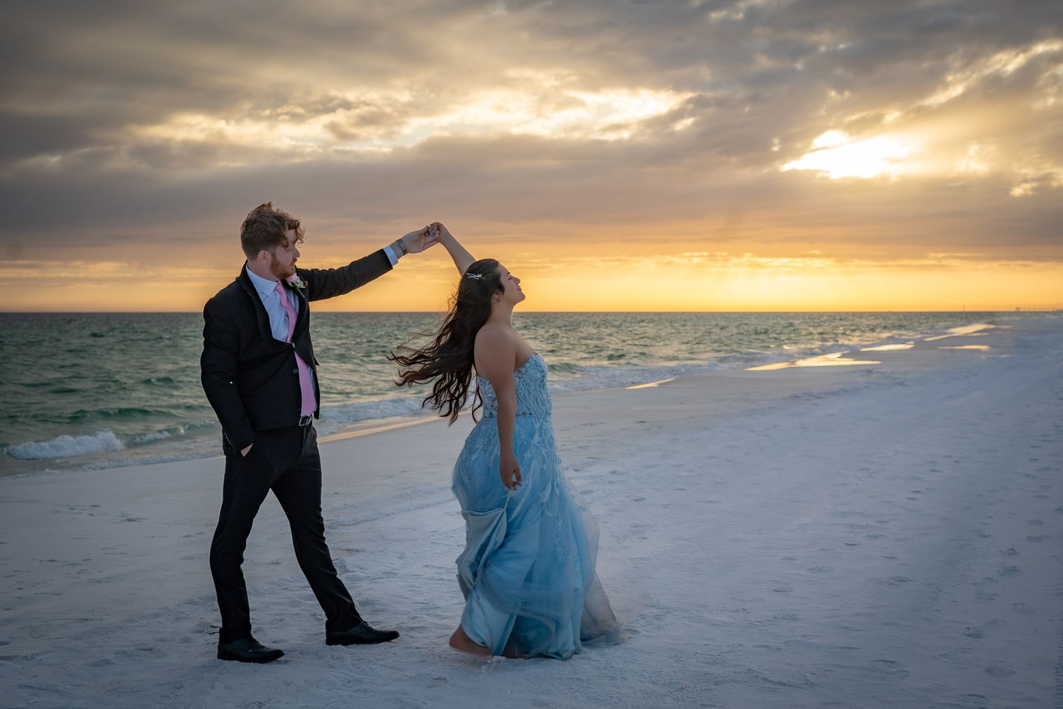 Couple dancing on a beach at sunset, man in black tuxedo spinning woman in flowing blue gown, dramatic cloudy sky.