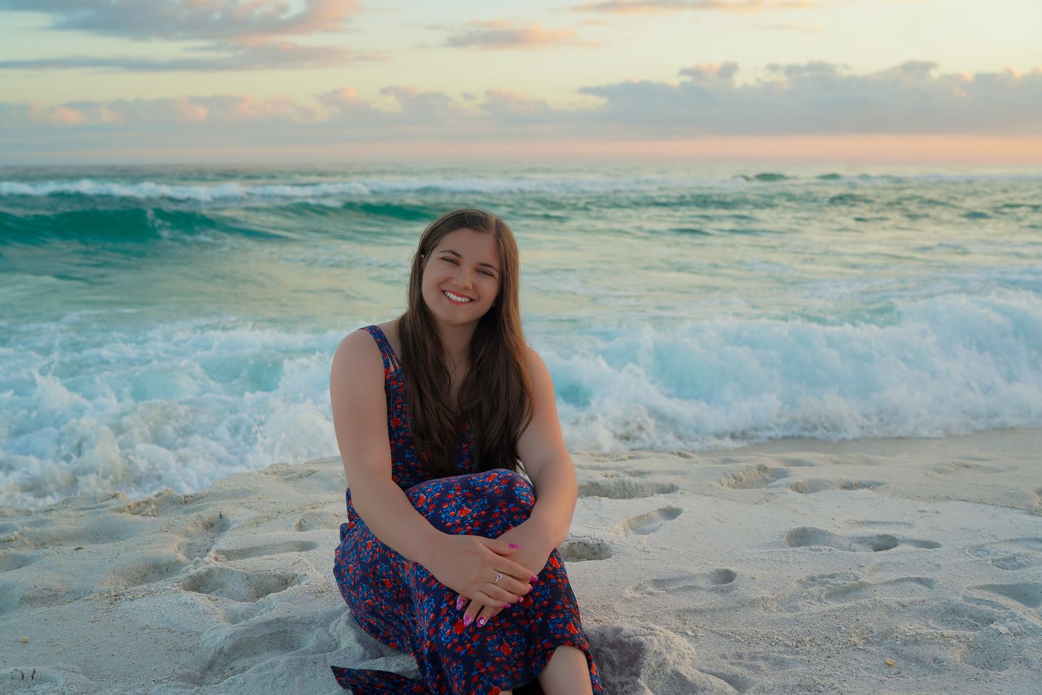 Smiling young woman in floral dress sitting on wet sand with crashing ocean waves behind.