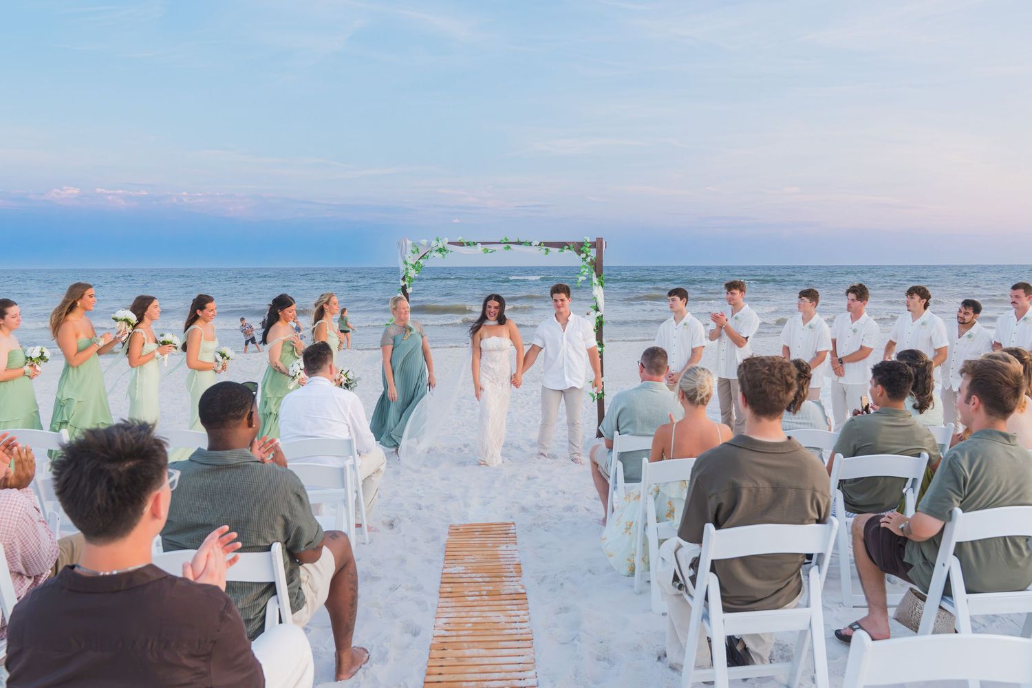 Beach wedding ceremony at dusk with bridal party at floral arch and seated guests.