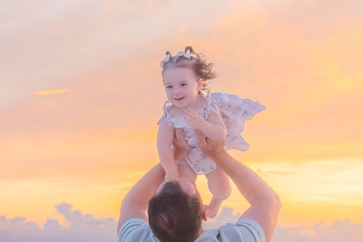 Father joyfully lifting smiling toddler girl in white dress against a golden sunset sky.