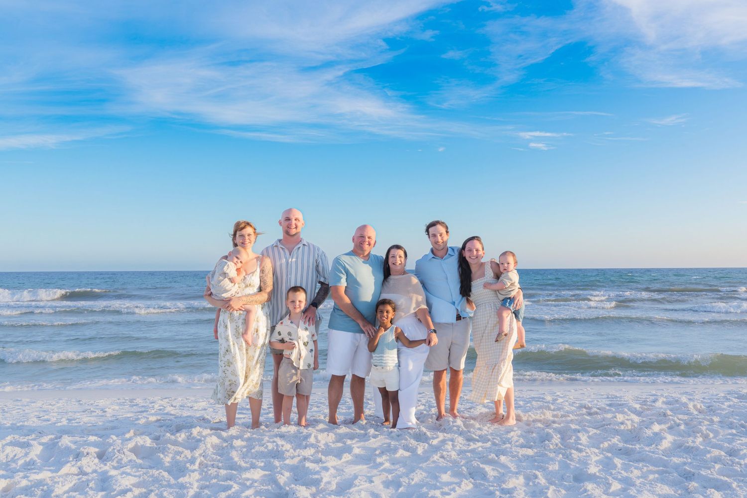 Multi-generational family posing together on a sunny beach with blue skies and ocean waves in the background.
