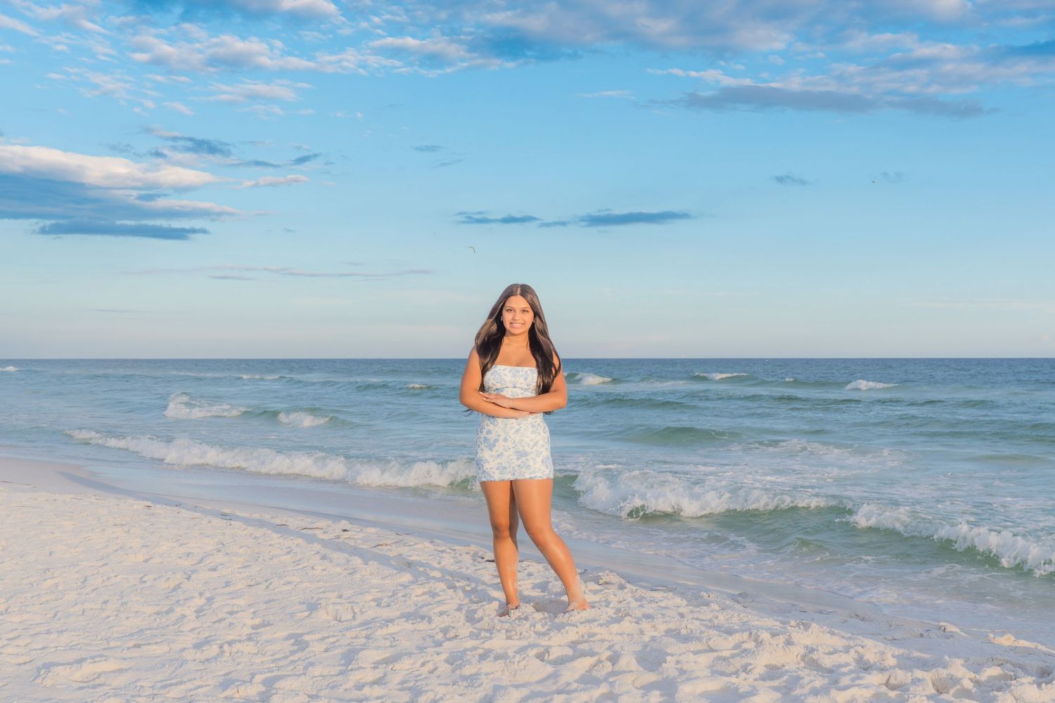 Young woman in white dress smiling on a white sandy beach with turquoise ocean waves and blue sky behind her.