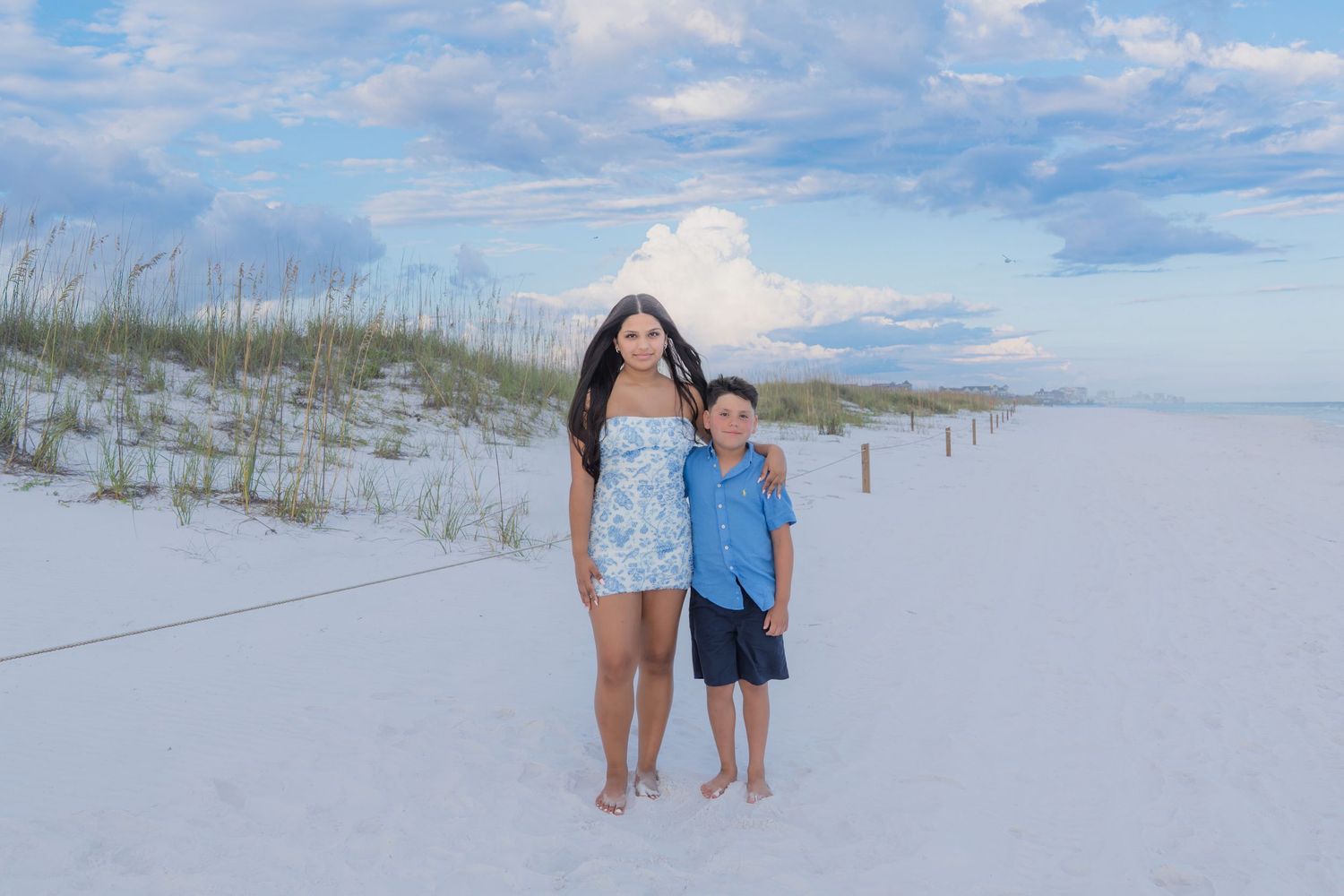 Woman and young boy posing together on white sandy beach with sand dunes and cloudy blue sky backdrop.
