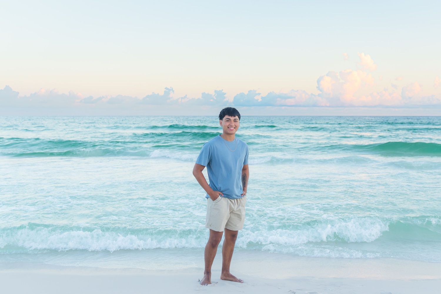 A young man smiles on a white sandy beach with turquoise ocean waves and a pastel sunset sky behind him.