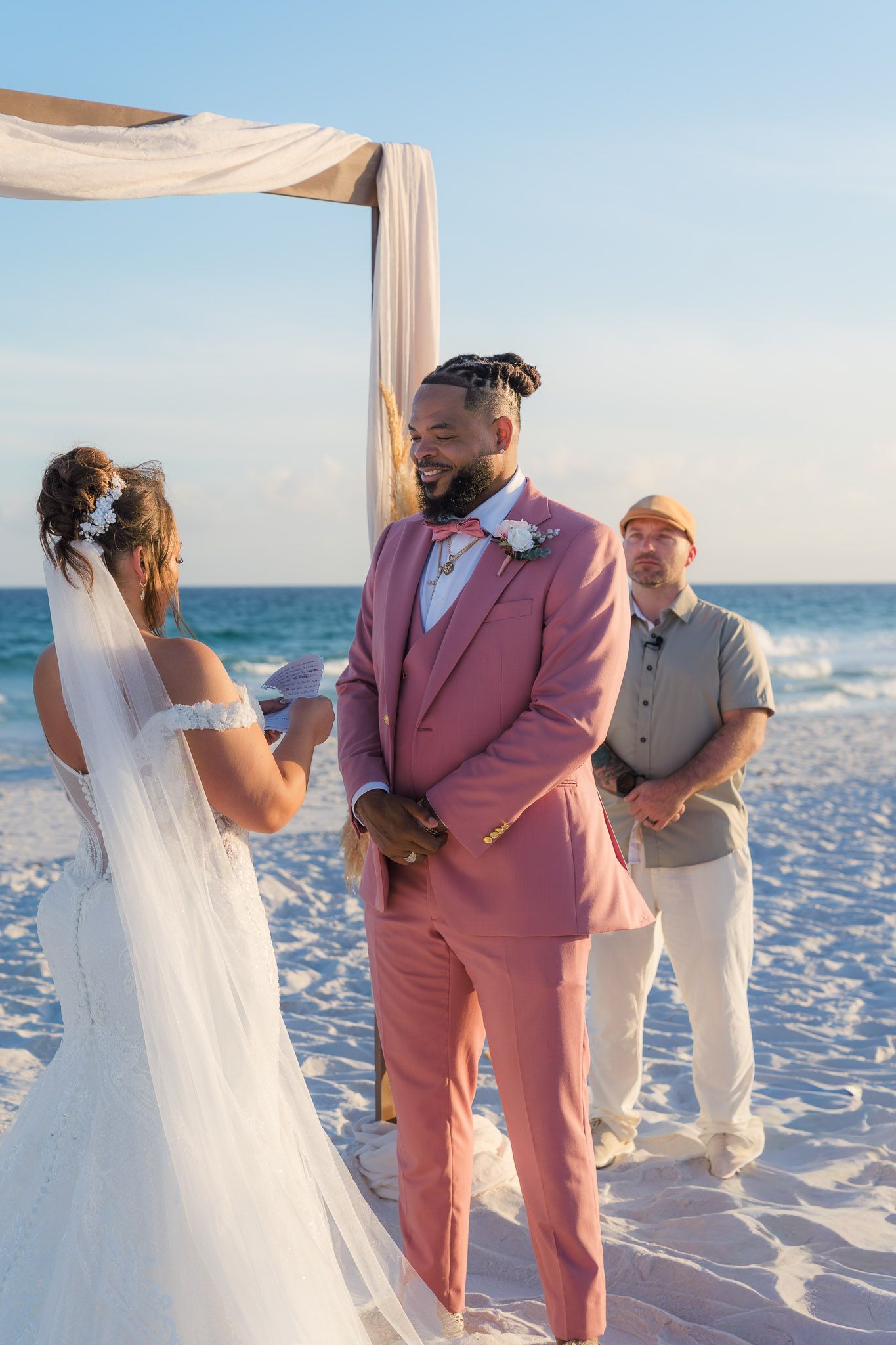 Groom in pink suit smiles at bride during beach wedding ceremony under draped arch with ocean backdrop.