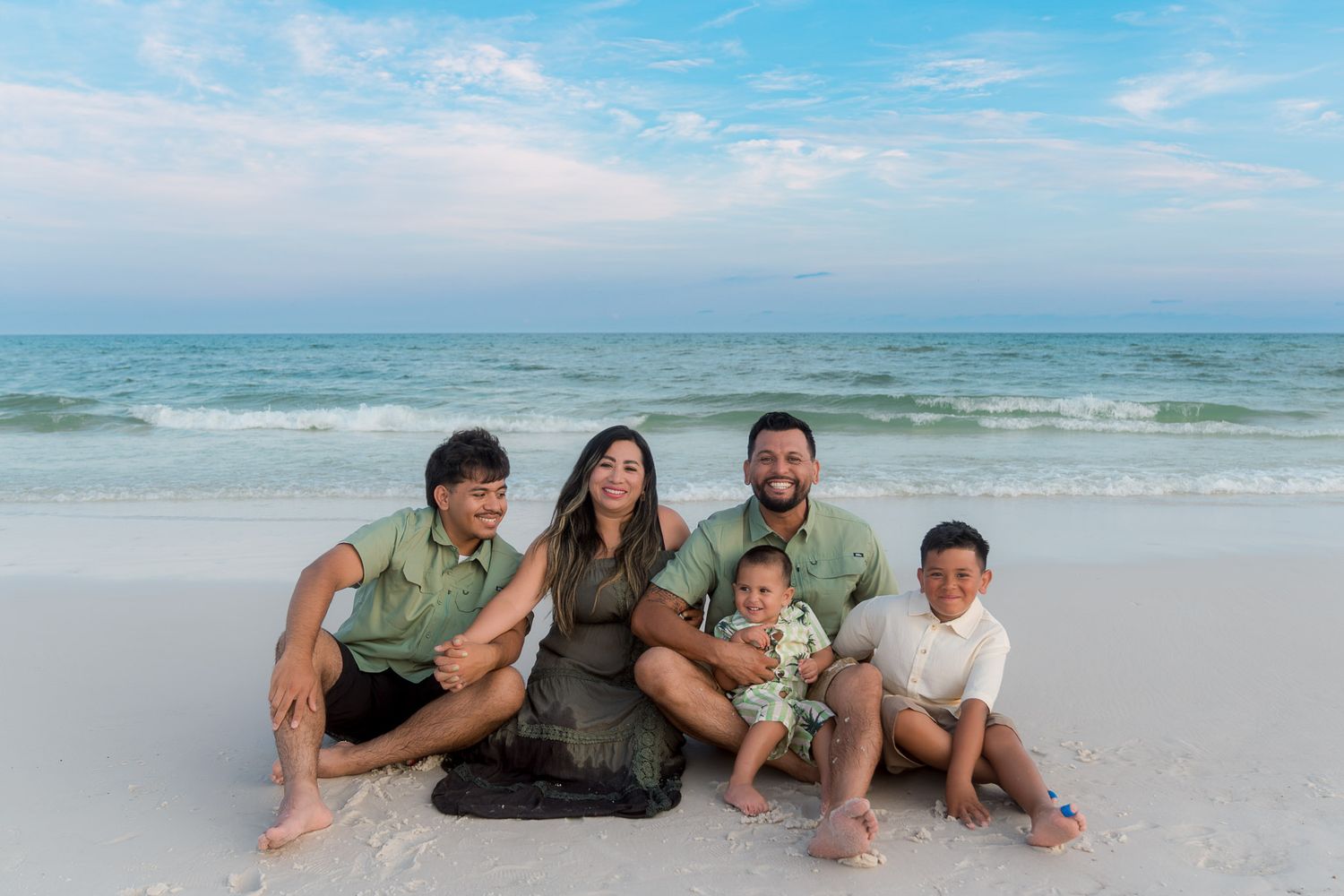 Family of five smiling together on a sandy beach with calm turquoise ocean waves and a pastel sunset sky.