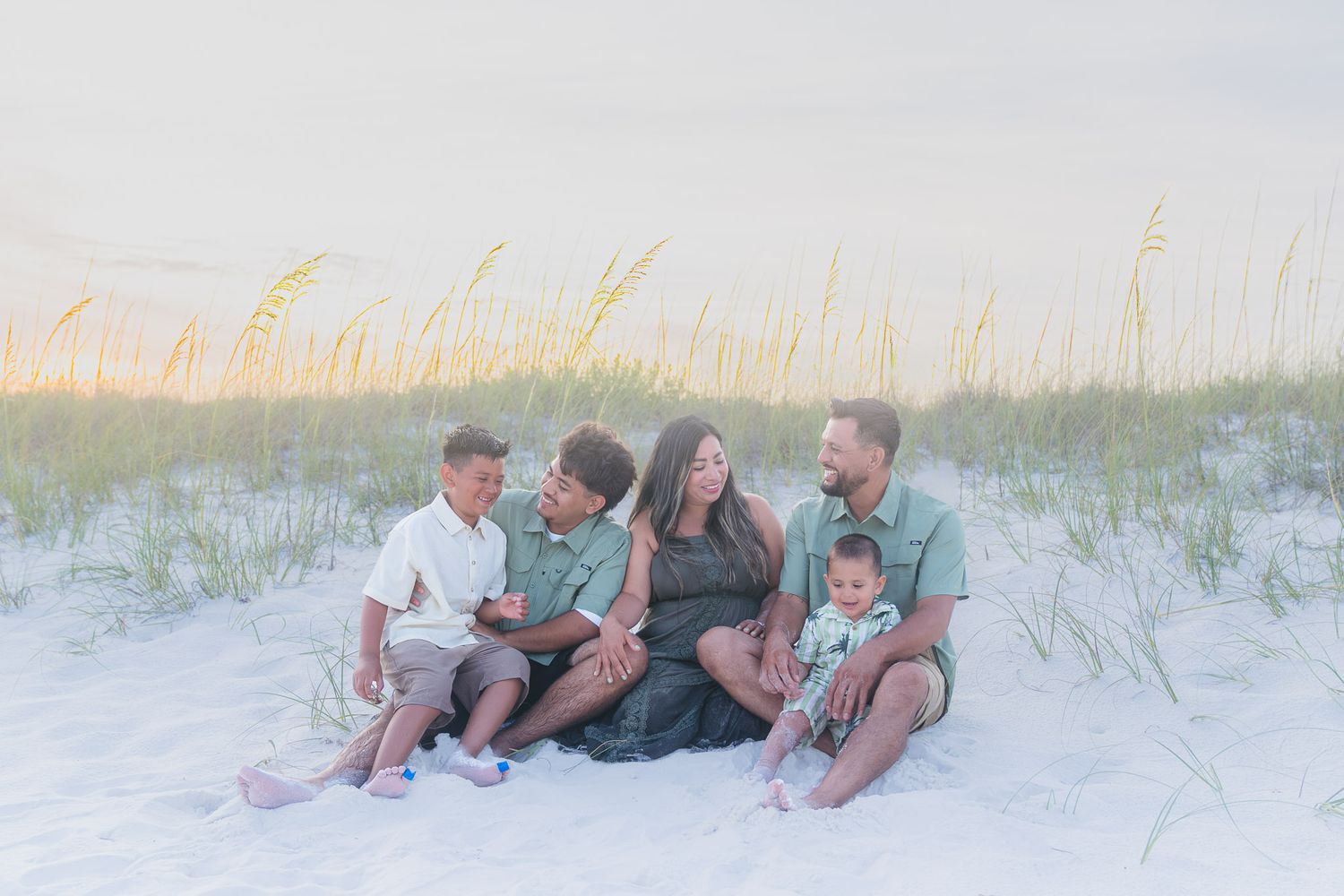 Family of five sitting together on white sand dunes with sea oats at golden hour.