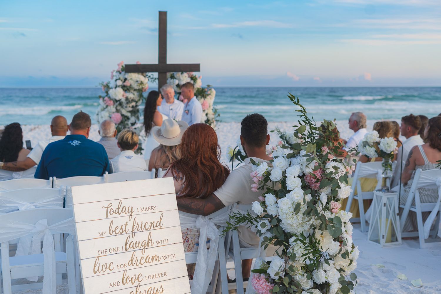 Guests seated at romantic beach wedding ceremony with floral arch cross altar and decorative sign.