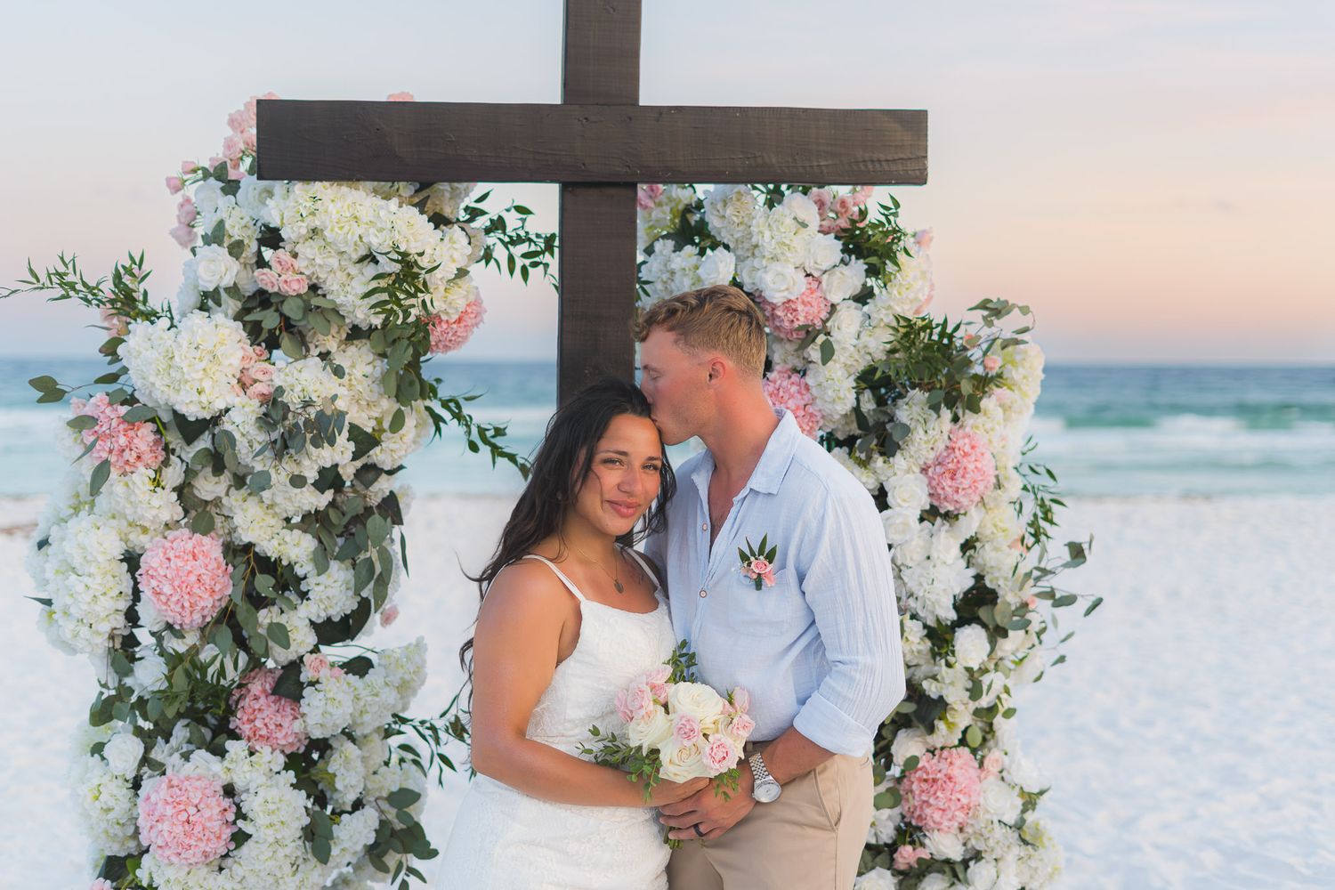 Couple shares romantic moment at beach wedding altar adorned with white and pink flowers and wooden cross at sunset.