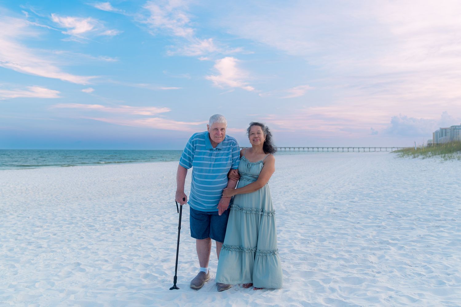 Couple posing on white sand beach at sunset, man with cane in blue polo, woman in teal dress, pier in background.