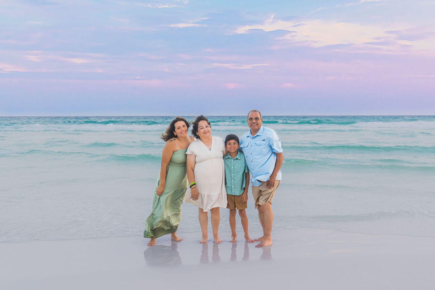 Family of four poses on a sandy beach at sunset with turquoise water and pink sky behind them.