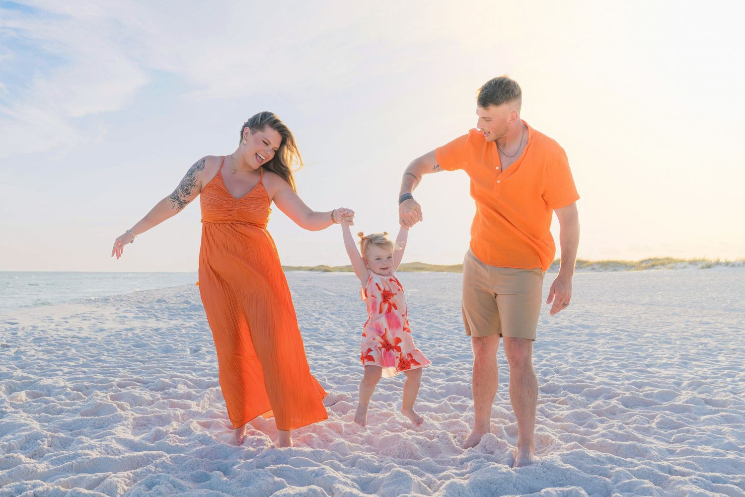 Family of three holding hands on a white sand beach, mom and dad in orange outfits swinging their toddler daughter.