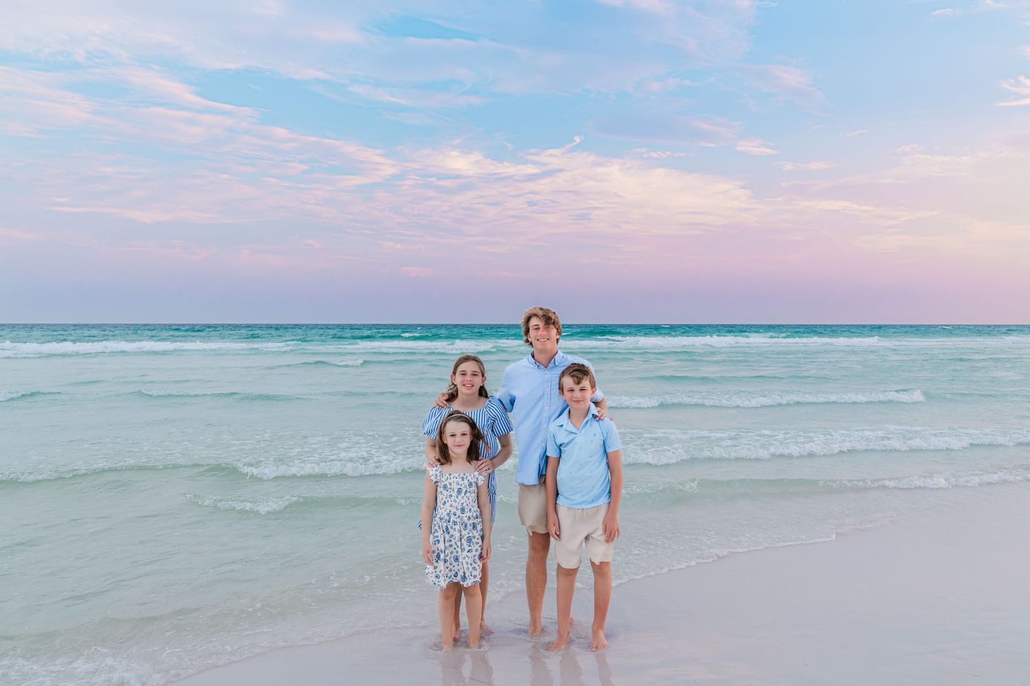 Family of four poses on a white sand beach at sunset with turquoise waves and pastel pink sky behind them.
