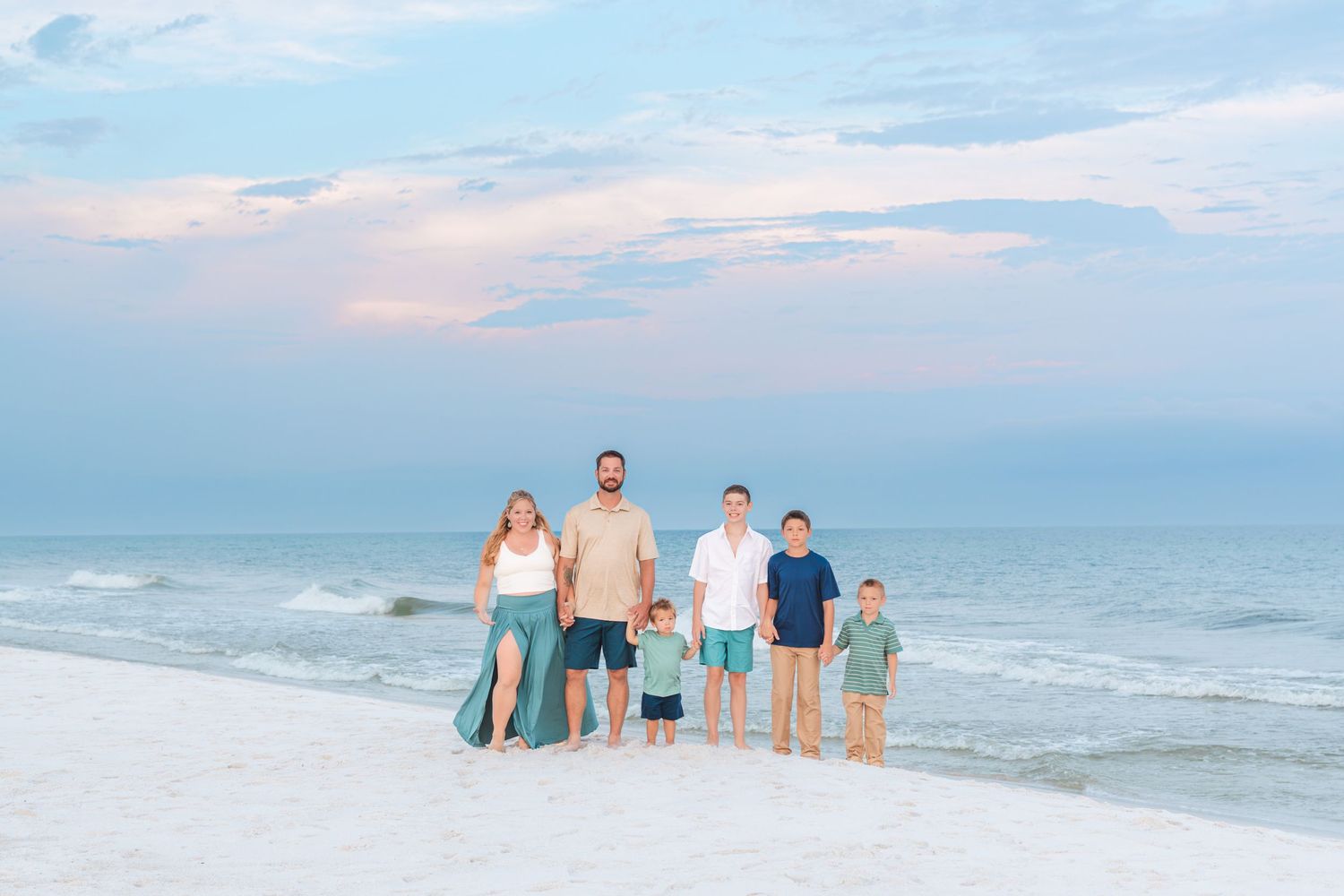 Family of seven posing on a white sand beach at sunset with calm ocean waves and pastel sky behind them.