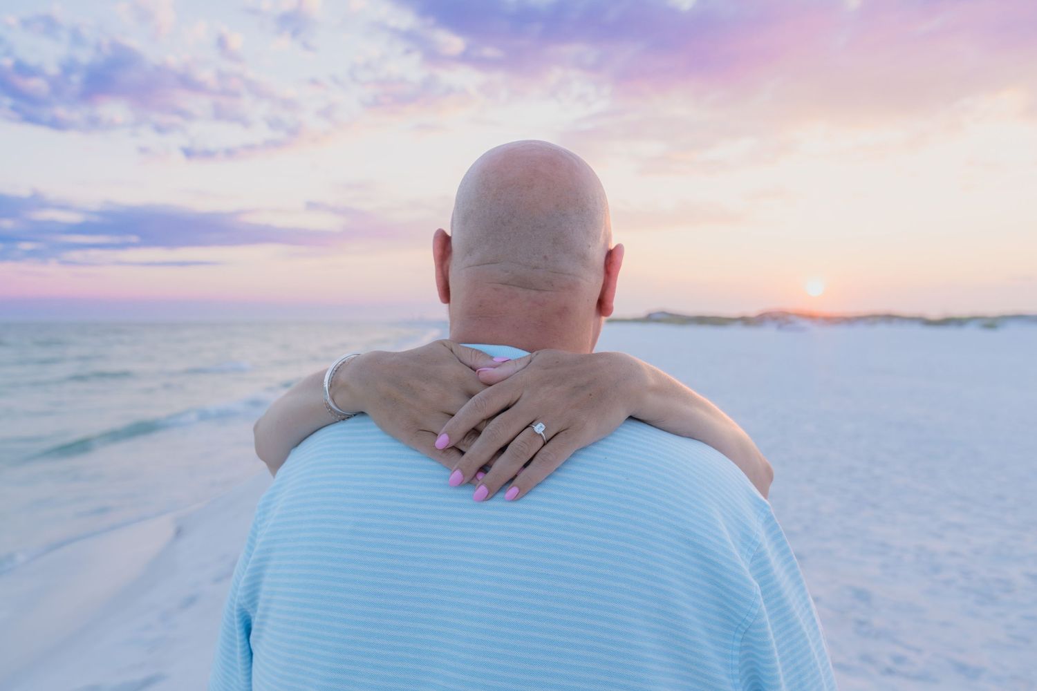 Couple embracing on a sandy beach at sunset, viewed from behind, with a pink and purple sky over the ocean.