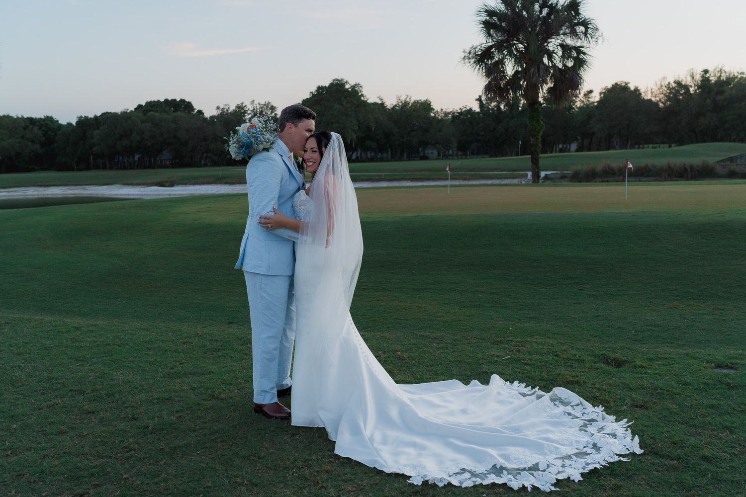 Bride and groom share a romantic kiss on a lush golf course at sunset, bride in elegant gown with long train.