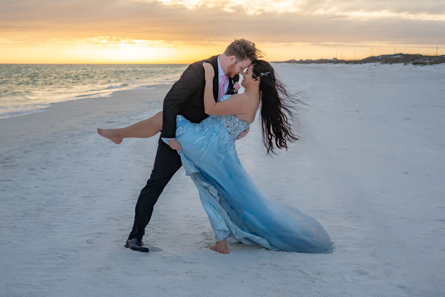 Groom lifting bride in a blue wedding gown during a romantic beach sunset photo session.