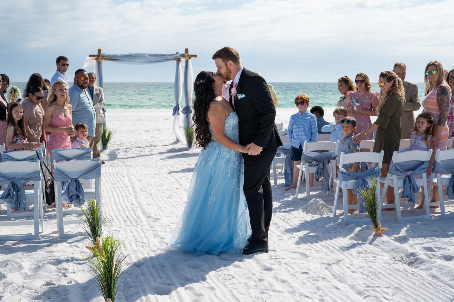 Bride in light blue ball gown kisses groom in black tuxedo at beach wedding ceremony.
