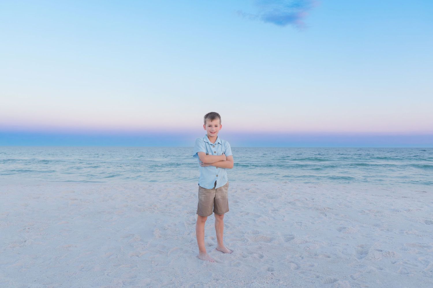 A young boy stands confidently with arms crossed on a white sand beach at dusk with calm turquoise water behind him.