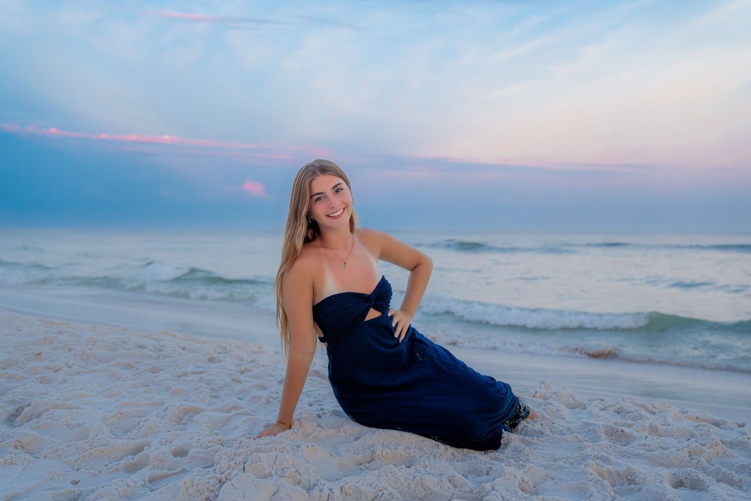 A smiling blonde woman in a navy blue dress poses on a sandy beach at sunset with ocean waves behind her.